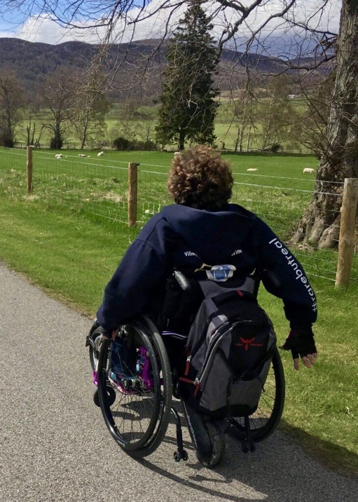 Photo of a person, with brown curly hair, using a self-propelling wheelchair on a countryside path. A large 'Phoenix Instinct Unstoppable' backpack is hung on the back of the wheelchair, below which a 'SmartDrive' power assist unit connected to the rear of the wheelchair can be seen. In the background a wire fence, fencing off a field in which a number of sheep, can be seen.