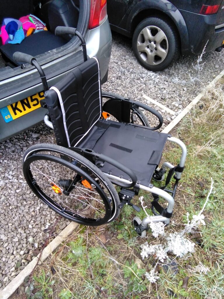 Photo of a 'Quickie Argon 2' rigid self-propelling wheelchair, by the open boot of a car next to an area of uneven ground covered in stones and small plants.