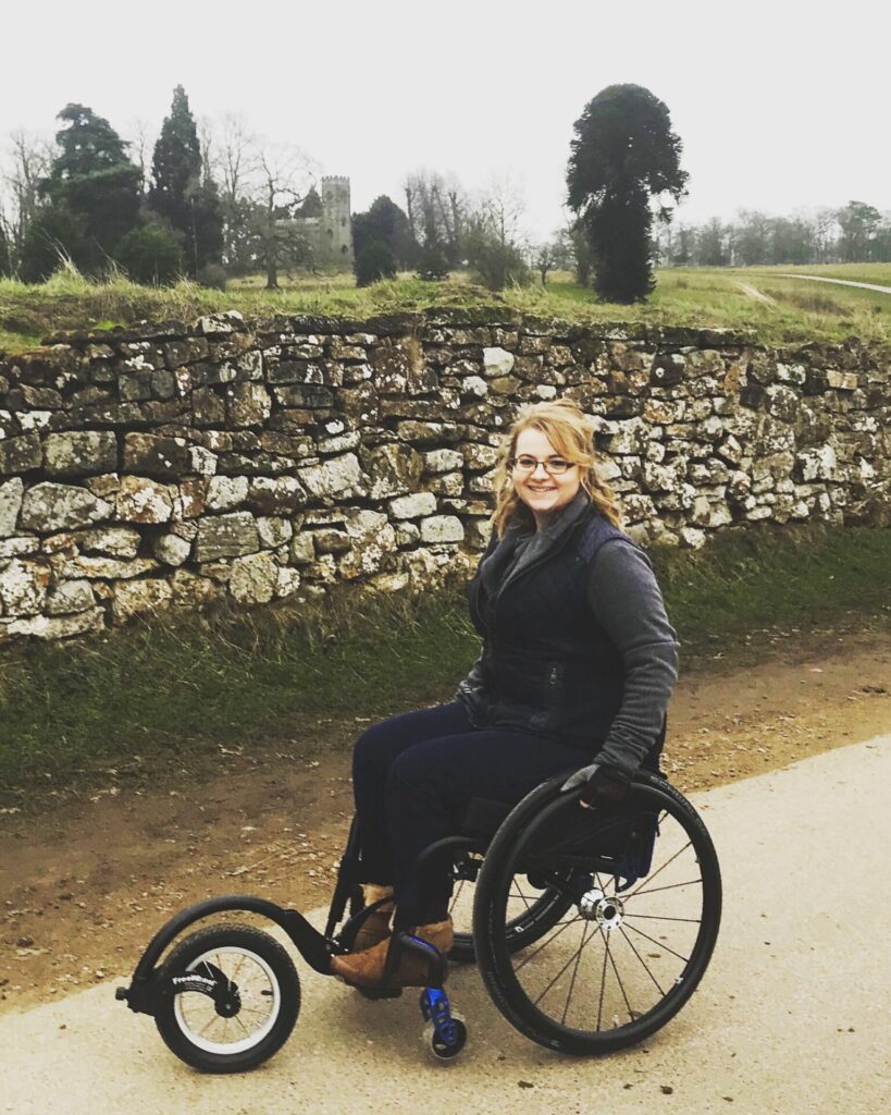 Photo of a person using a 'Quickie Helium' self-propelling wheelchair. A 'FreeWheel' attachment that is connected to the footrest, allows for additional manoeuvrability.