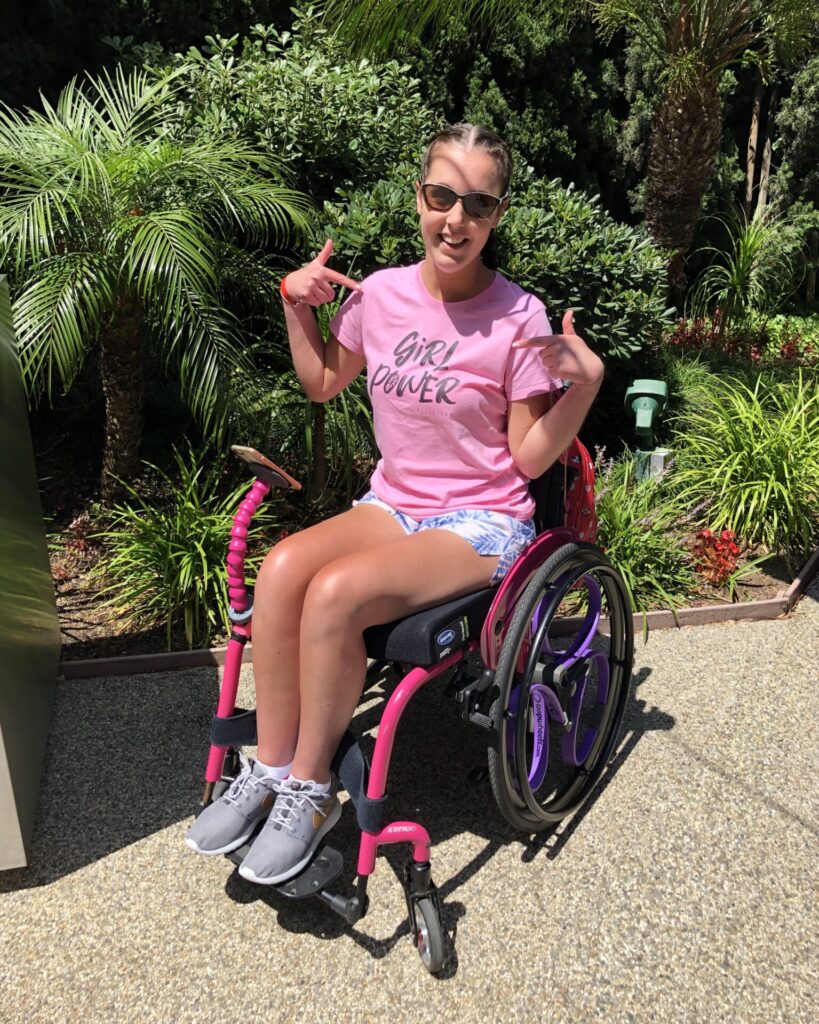 Photo of a woman wearing a pink shirt & shorts, using a pink 'Quickie Xenon' self-propelling wheelchair. The woman is pointing to the 'Girl Power' slogan emblazoned in black text on her shirt with both hands. Many tropical trees & plants fill up a garden plot in the background.