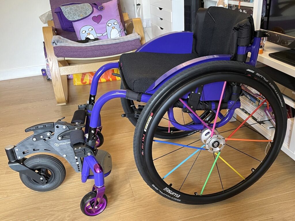 Side profile photo of a wheelchair with a 'RGK Frontwheel' attachment connected to the footrests.