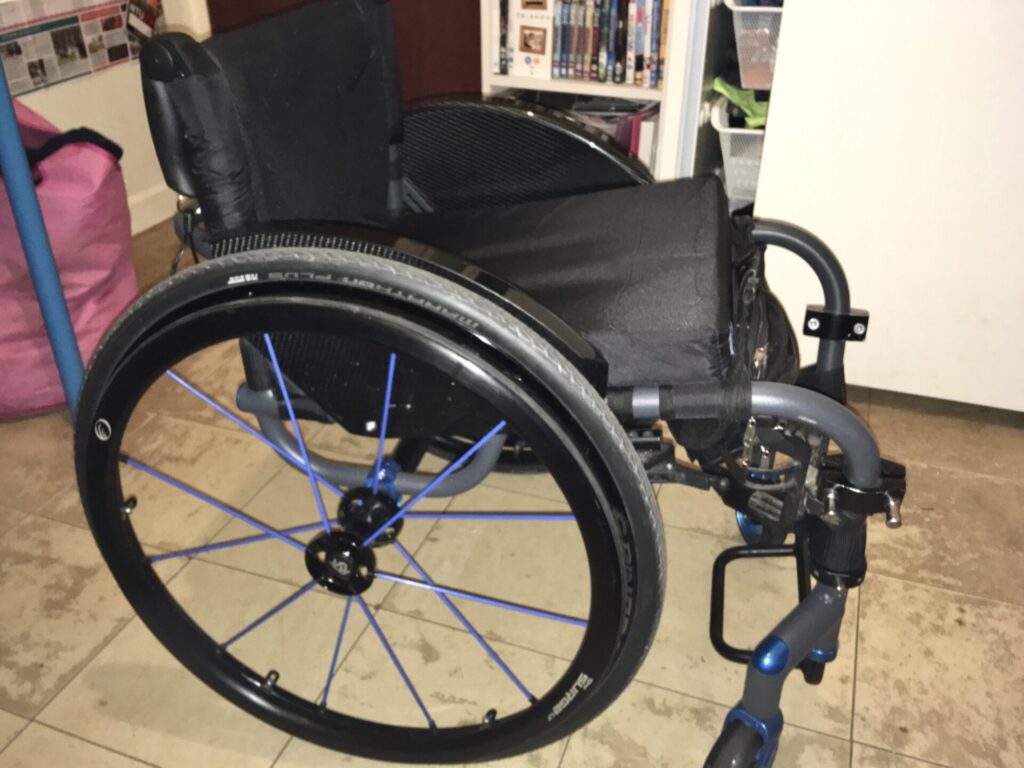 Side profile photo of an 'RGK Tiga' self-propelling wheelchair with blue highlights on the rear wheel's spokes.