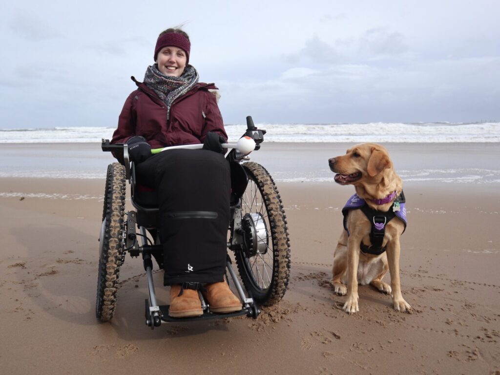 Photo of a female wheelchair user, wrapped up warm in a red beanie, scarf, red winter coat, & wheelchair parka, with her 'Canine partner' assistance dog (identified by their purple coat), posing for a photo at a beach, by the seafront. The woman is holding a dog ball launcher, to use to throw balls for her 'Canine partner' to fetch.