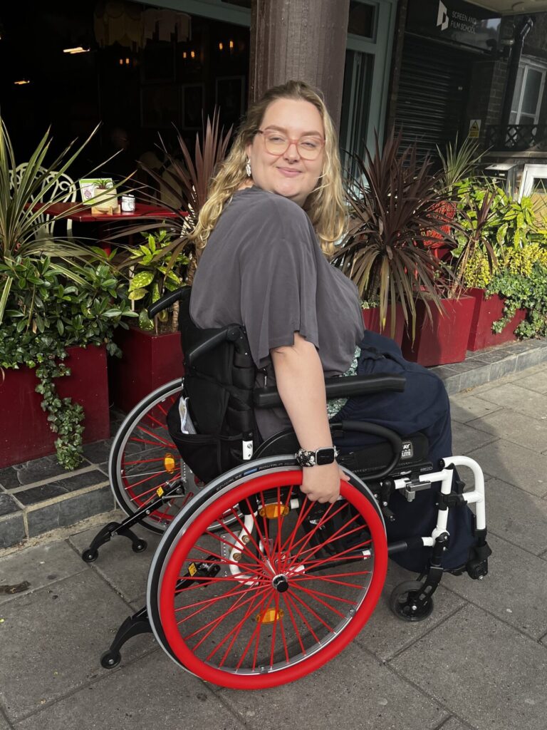 Photo of a blonde, bespectacled, female wheelchair user who has placed bright orange coloured 'RehaDesign Push Rim Covers' onto their wheelchair's push rims.