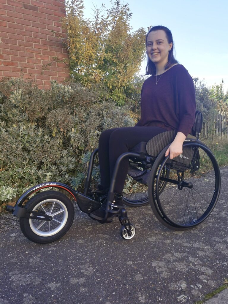 Side profile photo of a smiling woman posing for the camera, in a self-propelling wheelchair with a Rehasense Track Wheel' wheelchair attachment connected to the footrests.