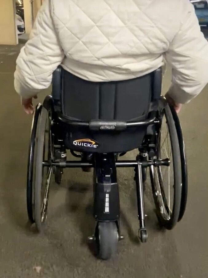 Back profile photo of a person using a 'Quickie' self-propelling wheelchair with a 'SMOOV One' rear power add-on connected to it.