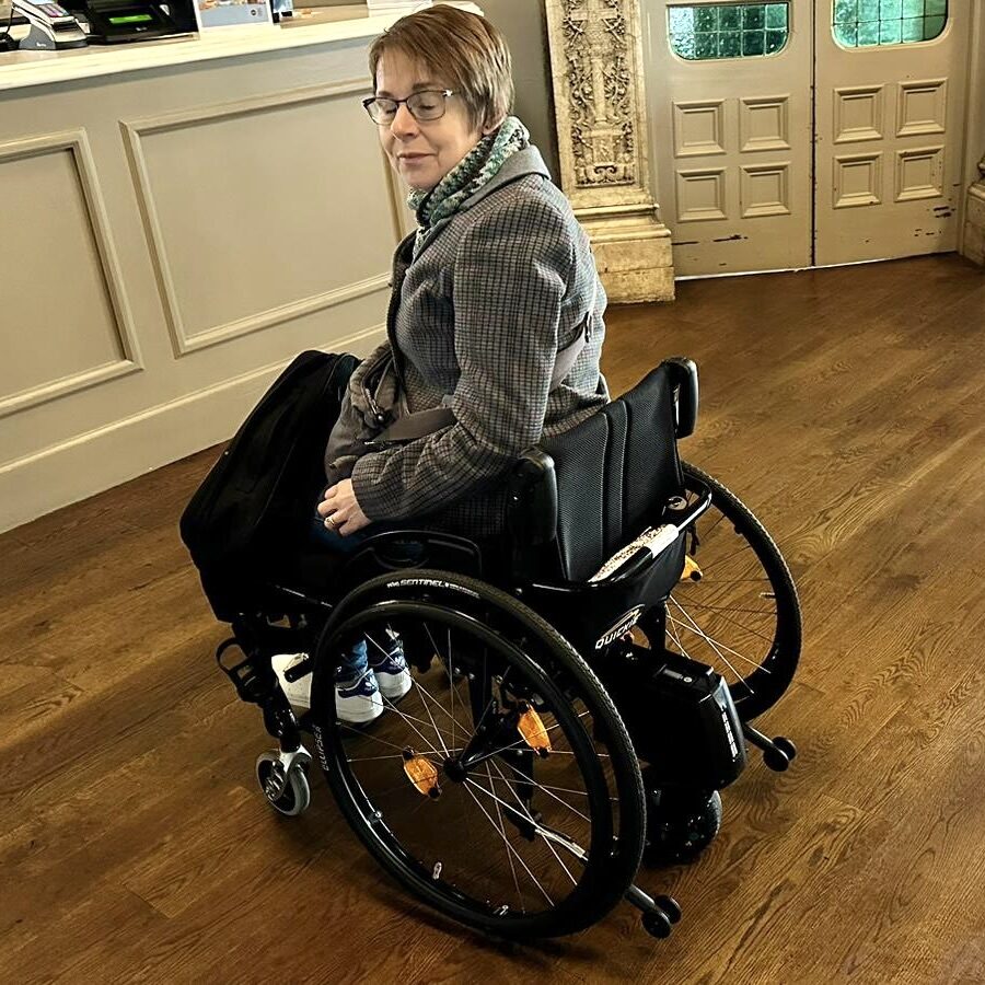Side profile photo of a woman using a self-propelling wheelchair with a 'SMOOV One' electric power-assist drive unit attached to the back of the wheelchair.