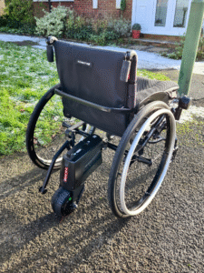 Back profile photo of a self-propelling wheelchair, showing the 'SMOOV One' power assist add-on, connected at the back of the wheelchair by an adapter axle, that then connects to the wheels.