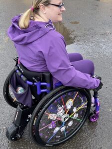 Side profile photo of a woman in a wheelchair with a 'SMOOV One' electric power-assist drive unit attached to the back of the wheelchair.
