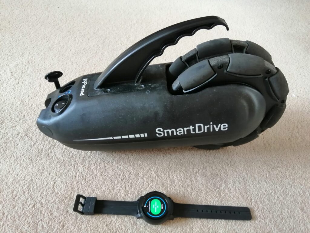 Top down photo of a 'SmartDrive MX2+' power add-on attachment for a manual wheelchair, & the smartwatch that controls it.