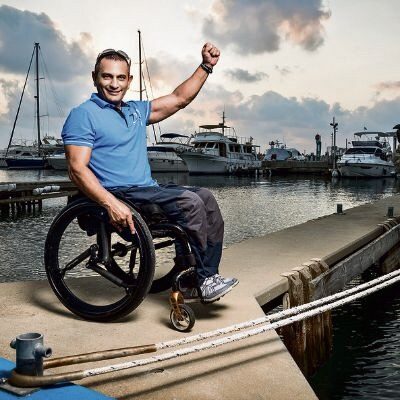 Photo of a man in a self-propelling wheelchair fitted with 'Softwheel' wheels, posing for a photo, with one arm raised in triumph, at a boat dock, with several large boats visible in the background.