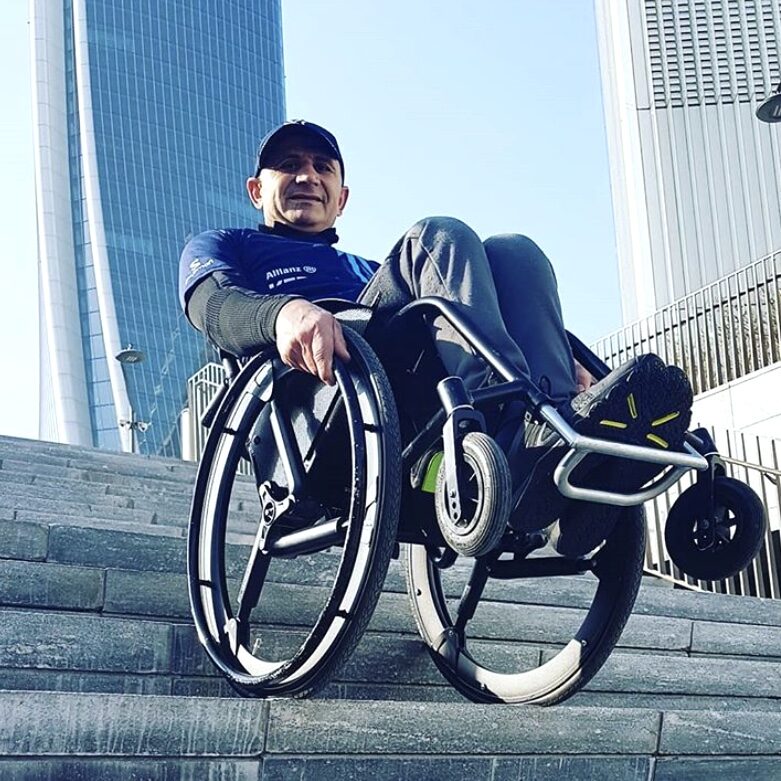 Photo of a man in a self-propelling wheelchair fitted with 'Softwheel' softshell model wheels, going down a set of steep stone stairs. In the background, several extremely tall buildings are visible.
