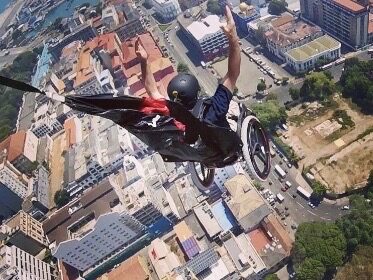 Photo of a man in wheelchair, wearing a helmet, a parachute on his back unfurling, as he is falling towards the ground in an urban city.