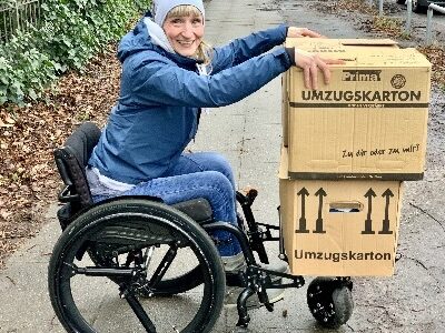 Photo of a woman using a self-propelling wheelchair with 'SoftWheel' suspension wheels installed onto it, & a front wheel attached, on top of which the woman is balancing two large boxes.