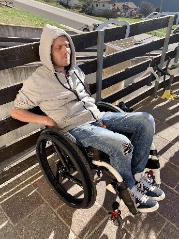 Photo of a man wearing a white/grey hoodie & jeans, resting against a guard rail made of dark wooden panels, while using a self-propelling wheelchair with 'Softwheel' suspension wheels, identifiable by the equidistant three suspension arms replacing regular wheel spokes.