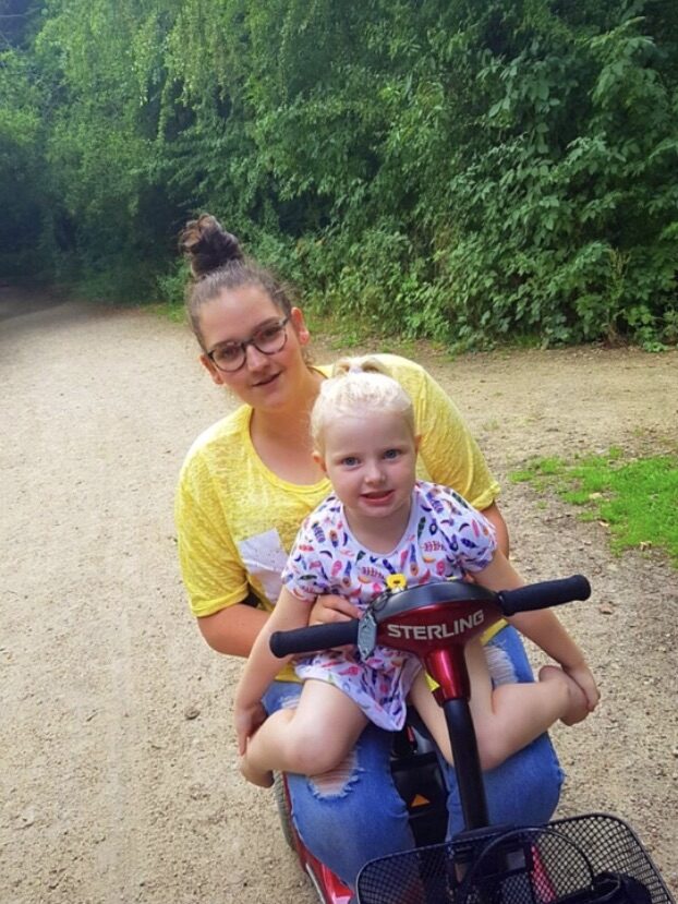 Photo of a woman using a 'Kymco Sterling Pearl' mobility scooter, on a rocky dirt path surrounded by large trees. The scooter is spacious enough to allow the woman to hold a small child on her while still having space to use the scooter, with the 'Sterling' branding visibly displayed in the centre of the scooters steering handles, directly in front of the young child.