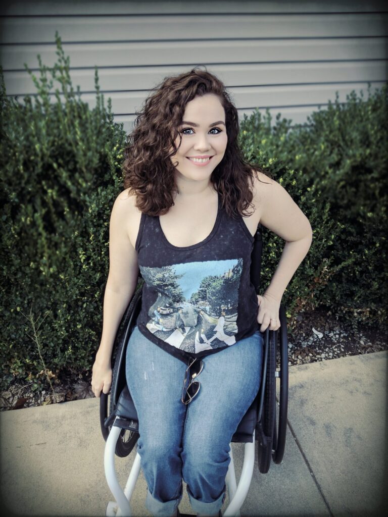 Photo of a smiling woman with shoulder length brown hair, using a 'TiLite Aero T' while posing for a photo in front of a row of plant shrubs running along the side of a building.