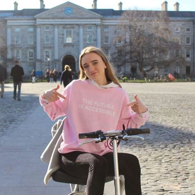 Photo of a young woman using a 'TravelScoot Deluxe' trike-style folding electric scooter, while posing for the photo in front of a large building, with both hands pointing with her index fingers at a white text slogan on the pink jumper she is wearing that reads 'The Future Is Accessible'