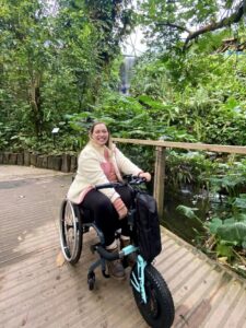 Photo of a woman using a self-propelling wheelchair with a 'Triride Compact HT' bike-style front power attachment connected, posing in front of a tropical plant display.