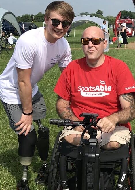 Photo two men, one older and one younger, both of whom are lower bilateral amputees. The younger man is wearing a white shirt and is leaning down slightly to pose for the photo using prosthetic legs. The older man is wearing a red 'SportsAble' shirt and using a self-propelling wheelchair with a 'Triride Compact Light' bike-style powered add-on attached.