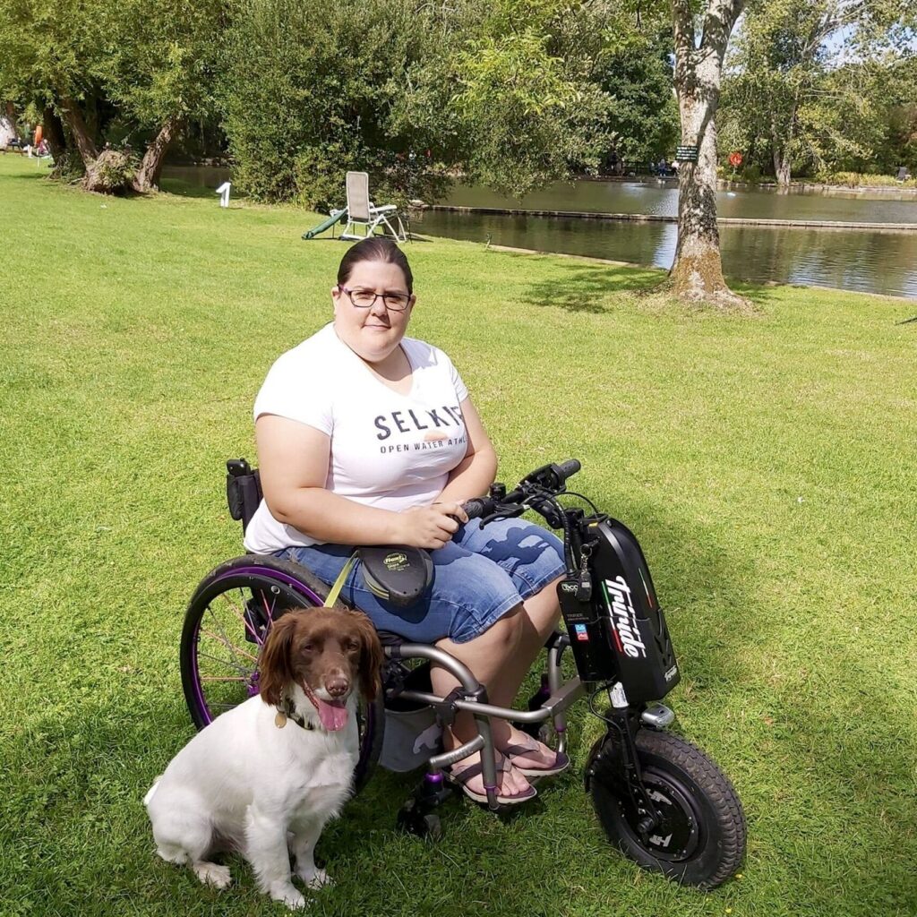 Photo of a woman, using a self-propelling wheelchair with a 'Triride Special Compact HT' bike-style power attachment connected at the front, on a day out in a public park.
