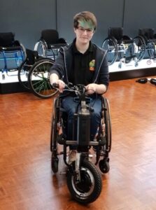 Photo of a person using a self-propelling wheelchair with a 'Triride Special Compact' power add-on connected. In the background, a number of wheelchairs can be seen on display.