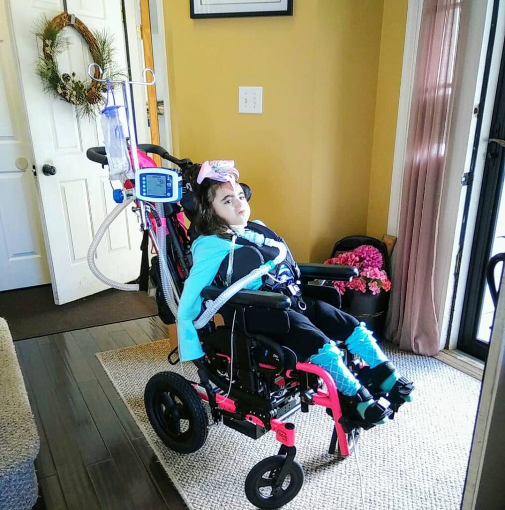 Photo of a young girl using a 'Zippie Iris' paediatric pink coloured wheelchair with a built-in tilt-in-space mechanism, chest harness, lateral support, straps, vent tray, activity tray, oxygen holder, and IV pole.