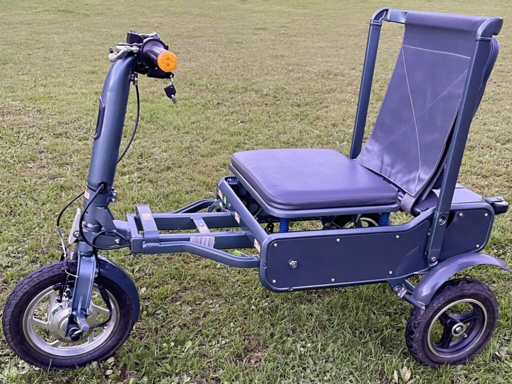 Side profile photo of an 'eFOLDi Explorer' trike-style mobility scooter in a grassy field, with its distinct blue colour on the frame, seat, & backrest.