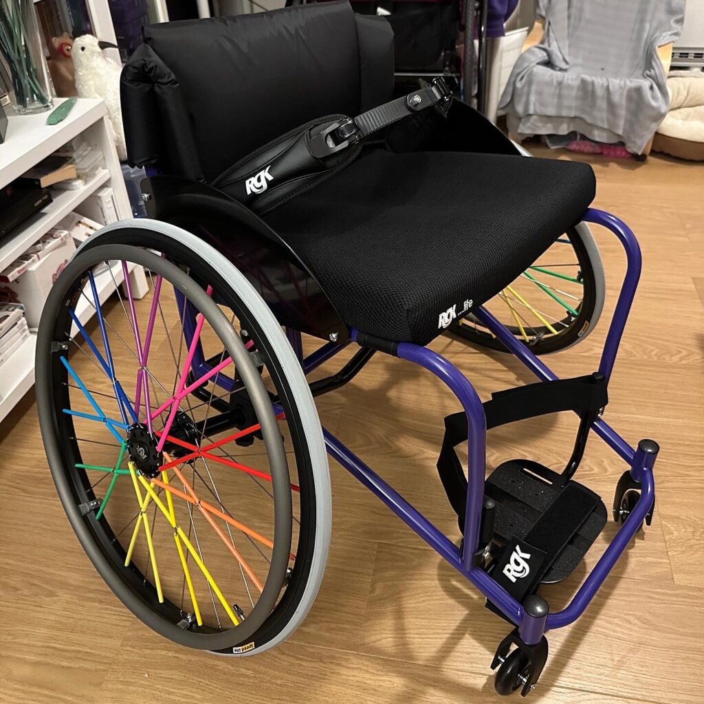 Side profile photo of a black 'RGK Club Sport' self-propelling wheelchair with multicoloured highlights on the main wheel's spokes.
