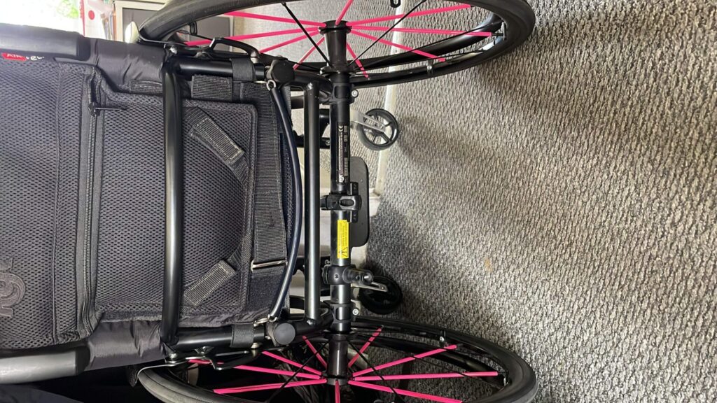 Back profile photo of an 'RGK Tiga' self-propelling wheelchair with pink highlights on the rear wheel's spokes and the 'RGK' logo on the top left of the back of wheelchair backrest.