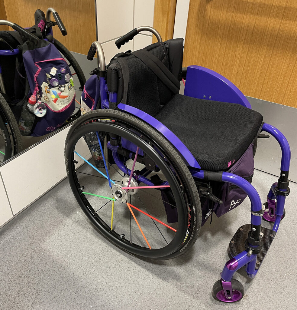 Side profile photo of a blue 'RGK Tiga FX' self-propelling wheelchair with multicoloured highlights on the main wheel's spokes.