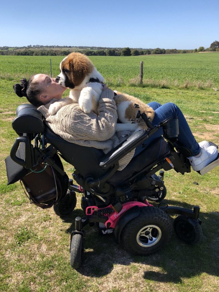 Side profile photo of a woman using a pink 'Frontier V6' all terrain electric wheelchair, reclined back at an angle, while hugging and showing affection to her dog, while out in the middle of a field.
