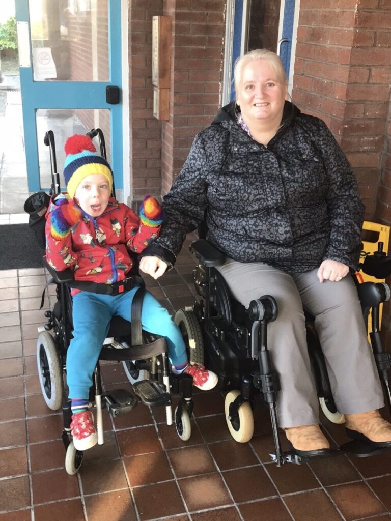 Photo of a young boy in a push chair, and an older woman using an 'Invacare Fox' powerchair, posing for a photo.