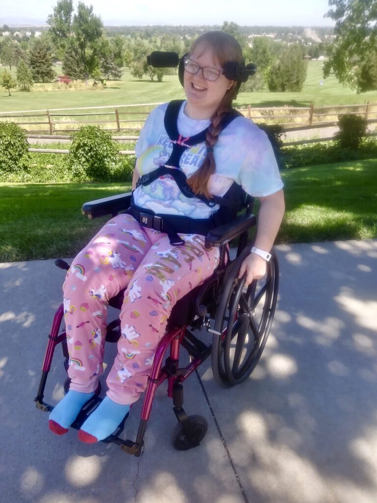 Photo of a bespectacled woman using a 'Kimobility Catalyst 5VX' folding wheelchair, on path of large slabs, overlooking the view of garden park.