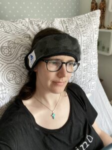 Photo of a bespectacled brunette woman propped up against a pillow on a bead, wearing a 'Koldtec Halo' headband- a wearable headband that enables you to apply ice to areas of your head as a method of cold therapy in order to relieve discomfort from a headache.