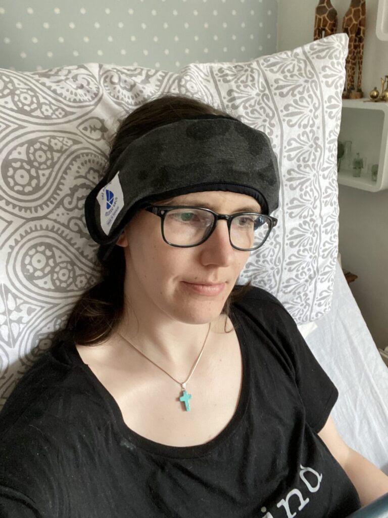 Photo of a bespectacled brunette woman propped up against a pillow on a bead, wearing a 'Koldtec Halo' headband- a wearable headband that enables you to apply ice to areas of your head as a method of cold therapy in order to relieve discomfort from a headache.