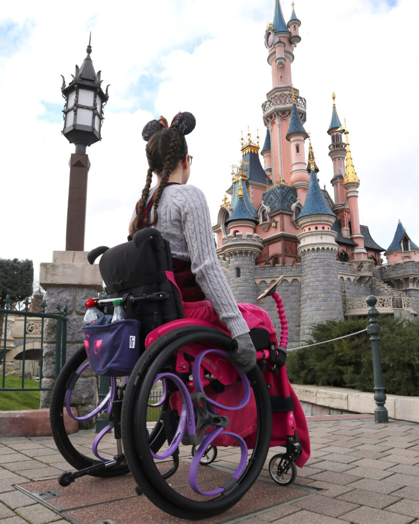 Photo of a woman using a self-propelling wheelchair in pink, with purple coloured 'Loopwheel Classic' wheelchair wheels. A theme park castle, in light pink but with blue & gold highlights, take up the majority of the background. As the woman is wearing a mouse ear shaped headband, it can be inferred that this photo was taken at 'Disneyland Paris'.