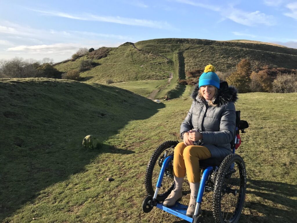 Photo of a woman using a 'Mountain E-Trike' in the middle of a grassy field surrounded by very hilly terrain. The variable elevation and uneven surfaces show that this Electric Trike is well equipped to cross uneven terrain.