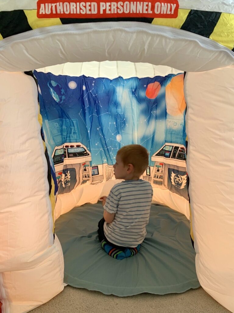 Photo of a young boy inside a 'POD's Play Sensory Tent' specifically the 'Galactic Space Adventure Pod' version, showing the children sitting down, surrounded by a 360° banner on that represents being onboard a space capsule, to help the children feel removed from the outside when in distress caused by sensory issues.