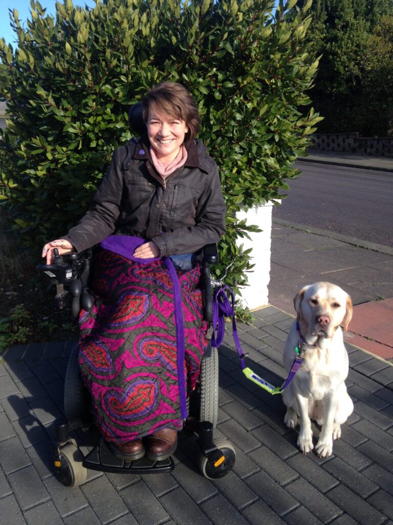 Smiling brunette woman using a 'Pride Quantum Q6 Edge' electric wheelchair, posing with a dog on a purple leash.