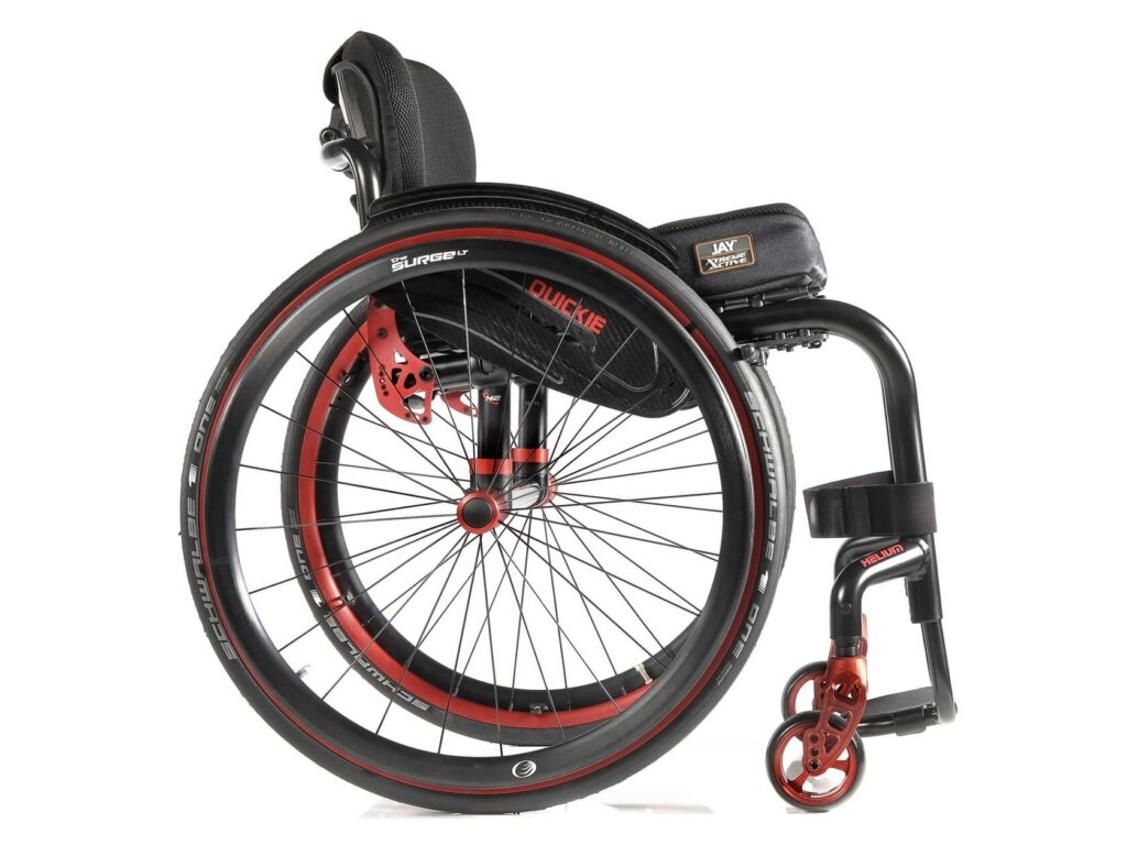 Side profile image of a 'Quickie Helium' self-propelling rigid wheelchair. Metallic orange highlights show off the main wheels, casters, & important fixings on the wheelchair frame.