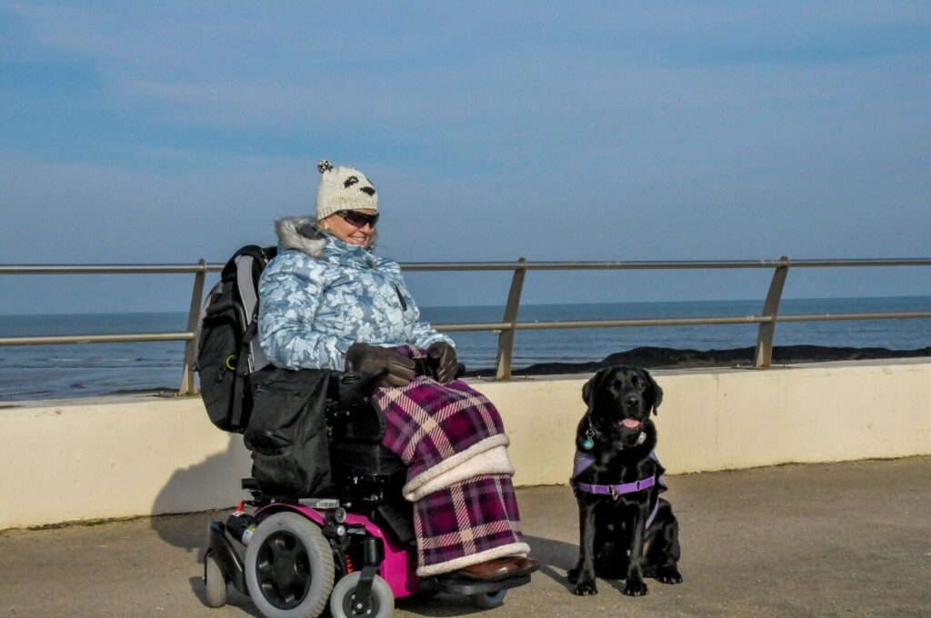 Photo of a woman using a 'Quickie Salsa M2' electric powerchair, wrapped up warm, while out on a day at the beach with a black labrador dog, posing for the photo on the beach promenade, overlooking a rocky beach with the sea stretching out to the horizon in the background.