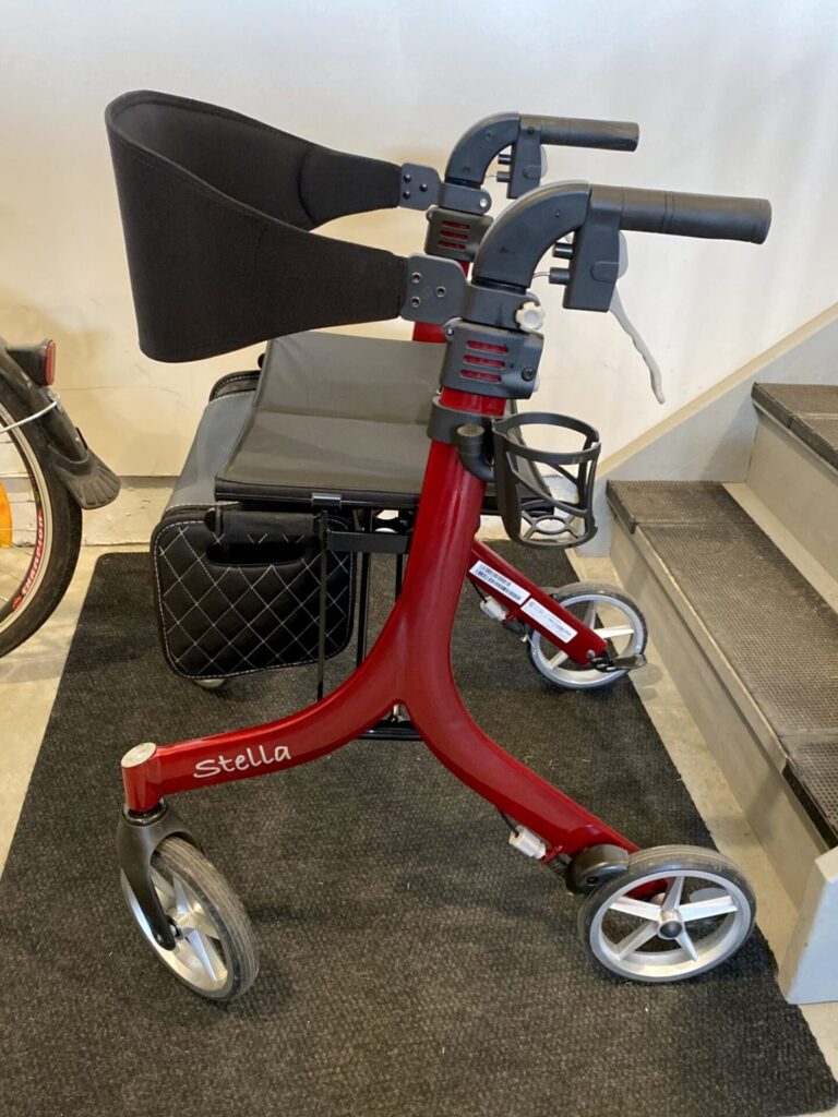 Side profile photo of a red coloured 'Stella Rollator' four wheel folding mobility aid, showing the supportive back strap, under-seat bag, and cupholder, as well as the comfort-moulded push handles with handbrakes.
