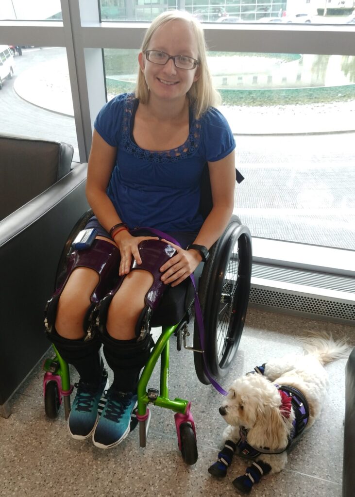 Photo of a smiling woman with shoulder length blonde hair, using a 'TiLite Aero T' while posing for a photo in front of a riverside window along with her small blonde haired dog.